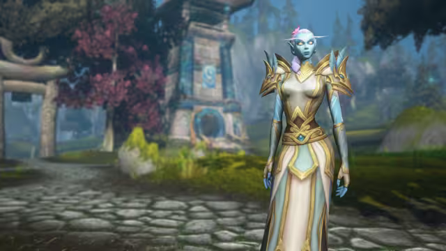 A blue-skinned woman with glowing orange eyes, long pointed ears and a lavender braid tucked with a pink flower stands in a floor-length cream, pale blue and gold robe with spiked gold shoulder guards. Out of focus behind her are a stone path, a tree with deep red leaves, a wooden arch and a stone tower marked with a blue swirl.