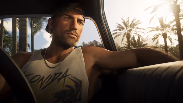 A bearded man in a backward cap, a chain necklace and a faded tank top printed with the word Leonida sits in a car with his arm stretched along the back of the bench seat, sunlight and palm trees outside the window.
