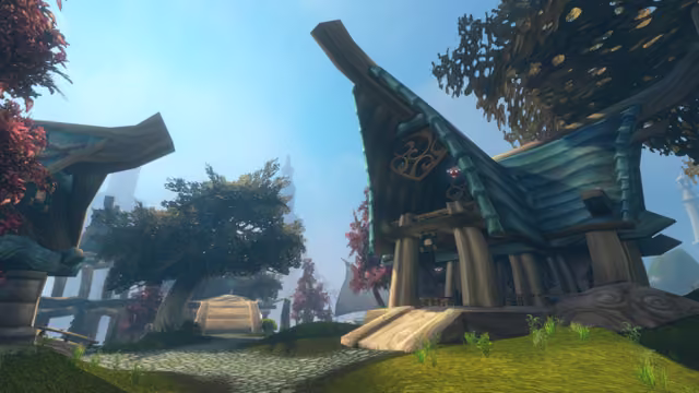 Seen from low on a grassy bank, a wooden building with a tall upswept roof of blue-green shingles, round carved ornaments and hanging lanterns stands on thick wooden posts. A cobbled path leads between it and another curved-roofed house toward wooden steps, a leafy tree and trees with red leaves, with pale towers faint in the blue haze.