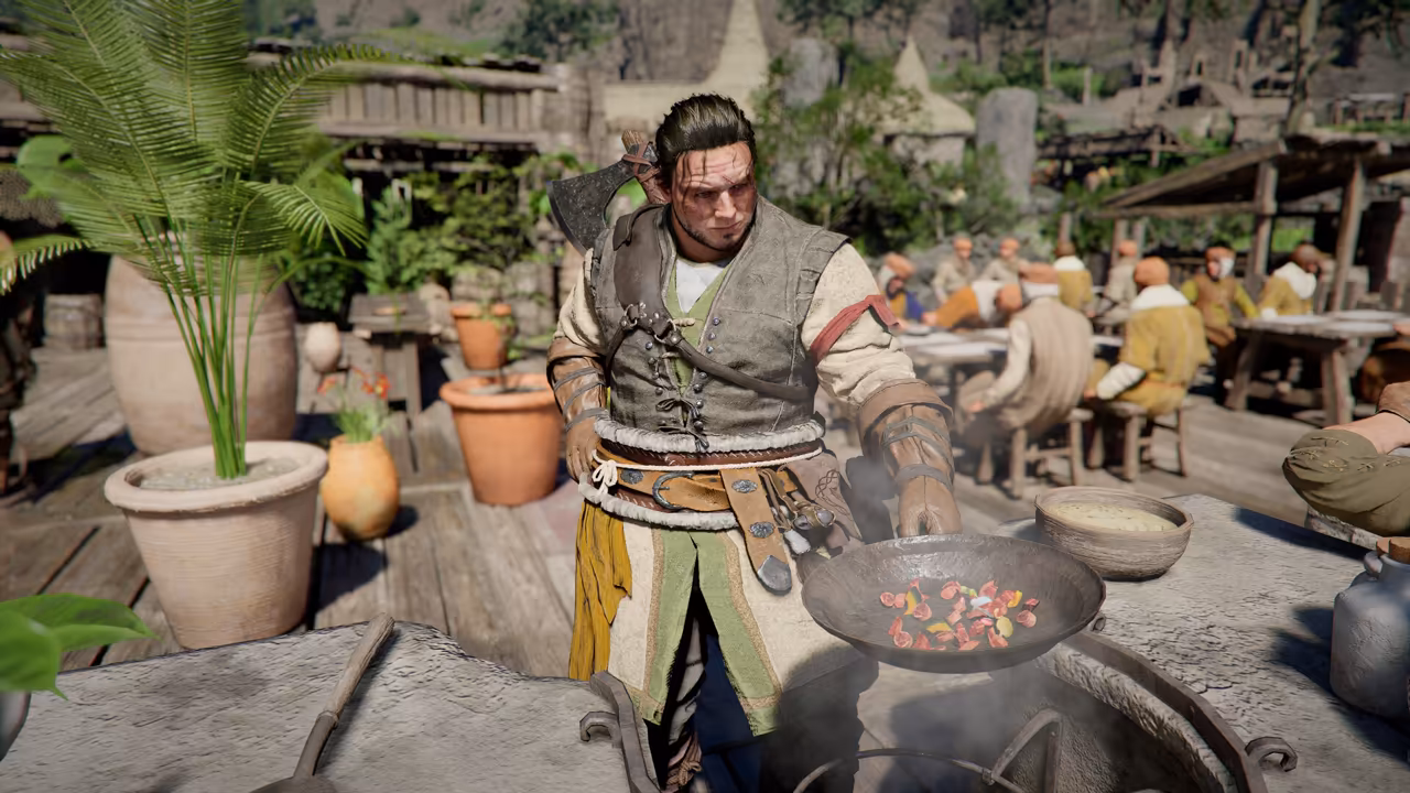 On a wooden terrace with potted plants, a man with slicked-back hair in a gray vest, rolled sleeves and a yellow apron holds a large pan of chopped red and yellow vegetables over a round stone oven, a bowl of sauce beside him. Behind him, people sit eating at long tables under awnings.