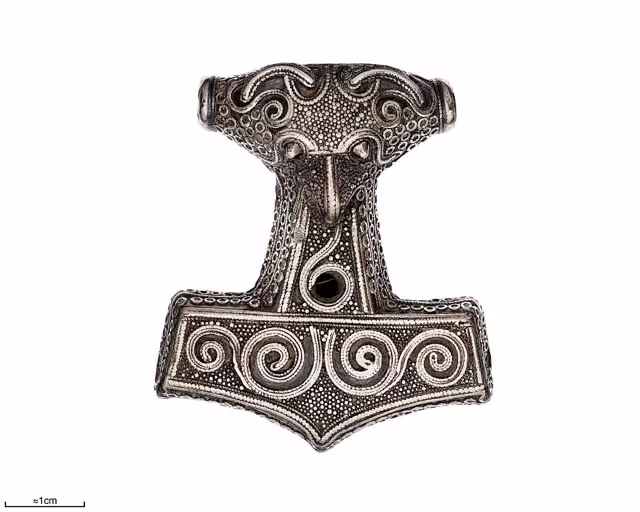 A silver hammer-shaped pendant decorated with filigree spirals and a stylised bird-like face on its upper part, photographed on a white background with a one-centimetre scale bar.