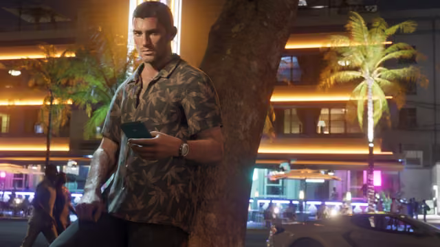 A man in a dark leaf-print shirt and a watch leans against a palm trunk looking at his phone, with glowing art deco hotels, outdoor tables and a sports car on the street behind him.