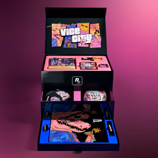 The black collector's box stands open on a magenta backdrop. The inside of the lid shows comic panels of an alligator, a lowrider, a sun in sunglasses, a flamingo and a manatee around the words Greetings from Vice City. A pink tray holds a boxed figure labeled Macca the Gator vinyl figure, a tin of Vice City art and two small boxes; a drawer below holds a black bag with Vice City patches and a white cap; the bottom drawer shows a large picture of a pink alligator in sunglasses raising a cocktail.