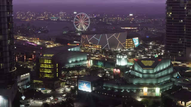 A high night view over a city: a large arena with a geometric light pattern, glowing cultural buildings and a billboard, busy roads, and a lit Ferris wheel by the bay under a purple sky.