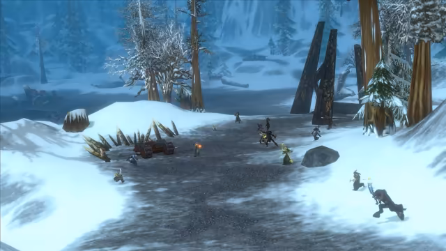 Seen from above, a dozen small figures in armor and robes fight with swords, staves and spells along a dark gravel road through deep snow. On the left, a wheeled wagon studded with spikes sits before a fence of sharpened stakes; snow-laden pines, tall bare trunks and a pair of leaning dark wooden pillars stand around a frozen pond behind them.