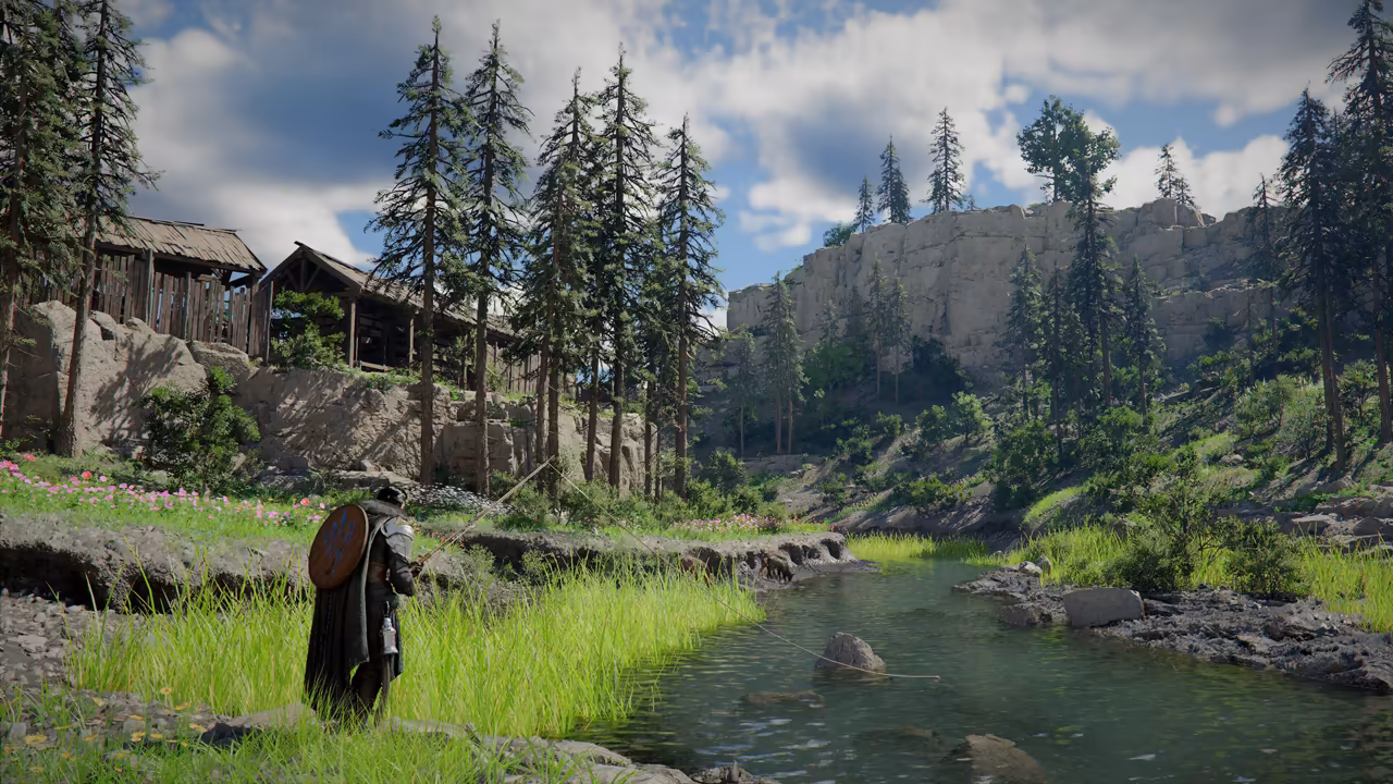Beside a clear, shallow stream running between rocks and bright green grass, a man in dark armor with a round shield on his back holds a fishing rod, its line reaching into the water. Wooden buildings sit on a rocky ledge at left, and pine trees and a pale cliff rise behind under a cloudy sky.