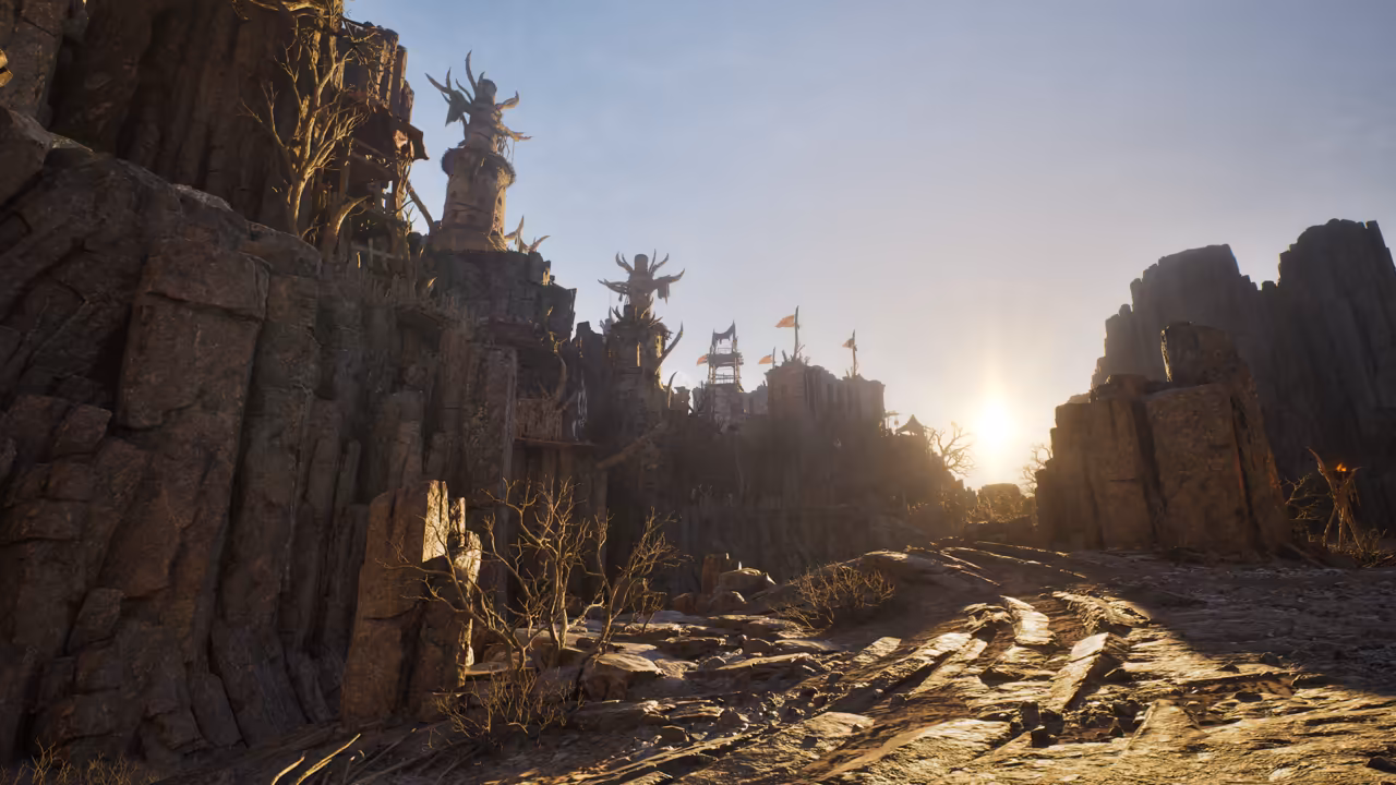 A dry canyon of columnar gray rock. On the clifftop at left stand towers topped with large spiked, horn-like sculptures, wooden scaffolds and red and orange flags. The rocky ground in the foreground is lit by a low sun shining between the cliffs at right, where a burning torch stands. Bare thorny bushes grow among the stones. No game interface is shown.