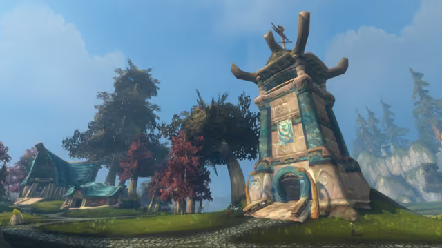 A tall tapering tower of pale stone and teal panels, crowned with upswept wooden beams and a small statue, stands on a grassy slope beside trees with red and green leaves. Houses with curling blue-green roofs sit along a cobbled path on the left, a few small figures stand near the tower's door, and gray cliffs with pines rise on the right under a blue sky.