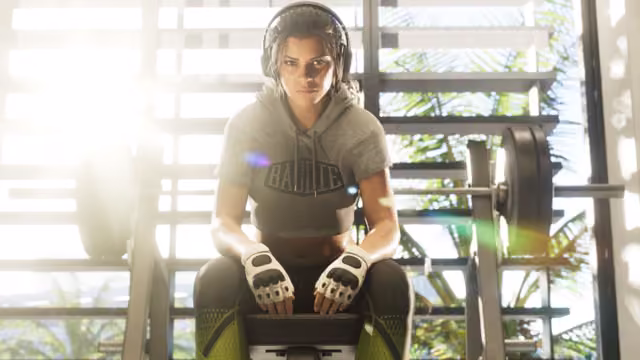 A woman wearing headphones, a gray cropped hoodie and white fingerless gloves sits on a bench in front of a barbell rack, bright light and palm leaves showing through the open steel frame behind her.