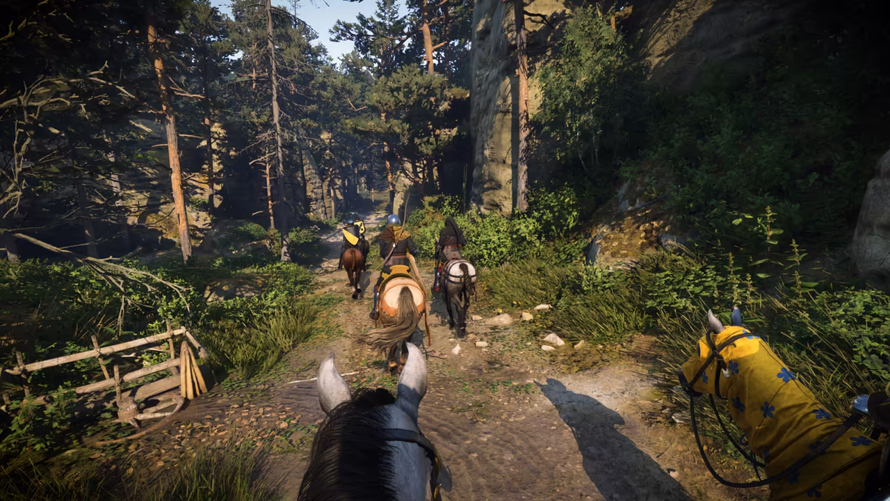 View between a horse's ears down a dirt path through pines, with three riders ahead and, at the right edge, a horse's head draped in yellow cloth with blue emblems.