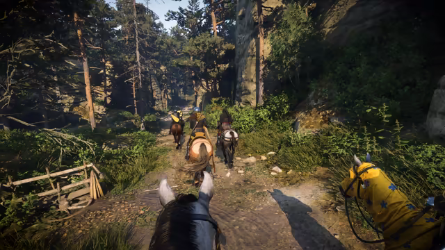 View between a horse's ears down a dirt path through pines, with three riders ahead and, at the right edge, a horse's head draped in yellow cloth with blue emblems.