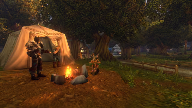 A campfire ringed with gray stones burns before a cream canvas tent under large trees, where a figure in dark red armor with a beast skull on one shoulder gestures toward it, a man in a wide-brimmed hat stands by the tent and a figure in leafy green and gold armor with an antlered helm kneels by the flames. Timber houses and a fenced road lie among the trees behind them.
