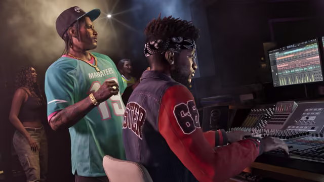 In a dim recording studio, a man with braids, a cap, gold chains and a teal Manatees football jersey watches a producer in a bandana and a varsity jacket work a large mixing console with waveforms on the screen, as two women stand in the background.