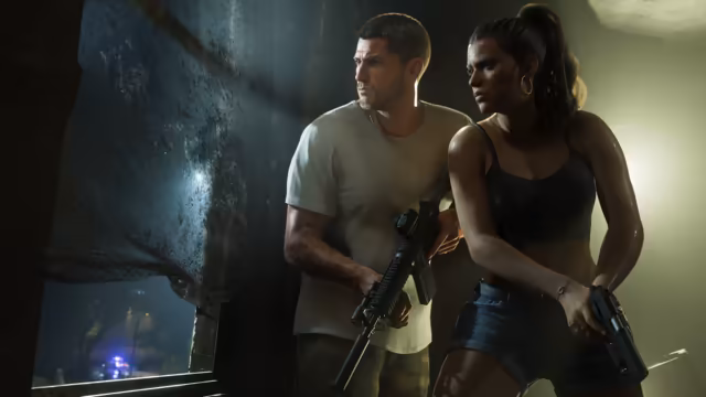 In a grimy, dim room, a man in a white T-shirt holding a rifle and a woman with a ponytail holding a pistol press against the wall beside a window and look out warily, a flashing police light faint outside.
