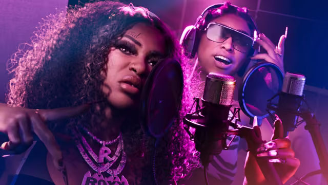 Two women perform into studio microphones with pop filters under purple light; the one in front has long curly hair, a small face tattoo and a diamond chain spelling Roxy, and the one behind wears headphones and jeweled glasses.