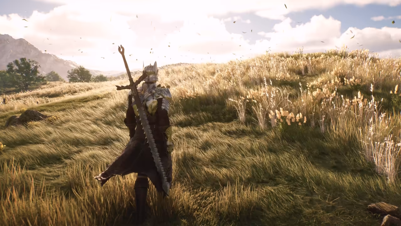 Bright daylight on a grassy hillside of tall pale grass and wildflowers bending in the wind, with leaves flying through the air. In the center, seen from behind, a figure in silver and gold armor and a long dark coat carries an enormous greatsword with a saw-toothed blade and a forked hilt across his back. Trees and a gray mountain stand at left under white clouds. No game interface is shown.