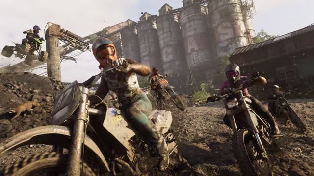 Mud-covered riders in helmets and goggles lean into a turn on dirt bikes along a muddy track, one airborne on a quad bike behind them and an animal running alongside, with graffiti-covered concrete silos and a rusted conveyor overhead.