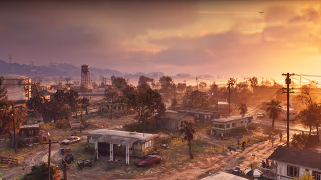 A high view of a run-down town under an orange and pink sky: mobile homes, sheds and dirt yards among palms and power lines, cars and quad bikes on the tracks, and a water tower and industrial buildings on the hazy horizon.