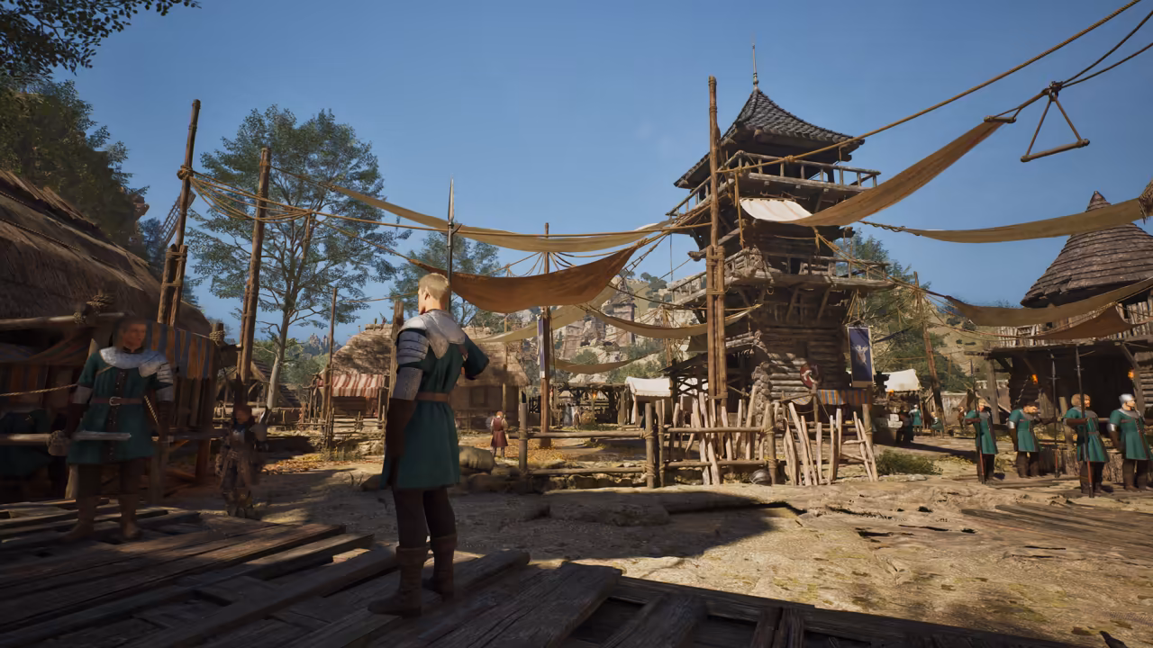 Bright daylight over a camp of wooden buildings. A tall log watchtower with a curved tiled roof rises at center, and sun-bleached canvas sheets hang from ropes between tall poles. A blond man in a green tabard over mail stands on a plank floor in the foreground; another guard in green stands at left, and a row of guards in green holding spears waits at right beside thatched huts. Trees and rocky hills stand behind. No game interface is shown.