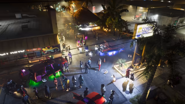 Seen from above at night, dozens of people stand and talk in a parking lot beside a thatched-roof bar and a shop painted with a gecko. A purple SUV with its doors open glows green underneath, a maroon car with its hood up glows blue, a red car sits with its doors open in the foreground, traffic cones dot the lot, and a lit sign for Conch King Seafood Kitchen stands among palm trees under power lines.