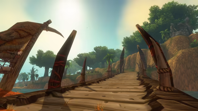 A low view along weathered wooden planks flanked by tall pale tusks and dark posts wrapped in red cord, with a hide shelter on the left. Ahead, broad-leaved trees and palms cover sandy hills, and a ruined stone structure sits on the green slope at right beneath a bright, hazy sky.