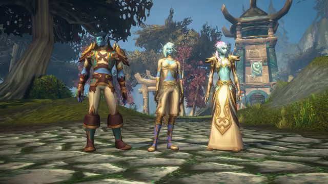Three blue-skinned characters with long pointed ears stand side by side on a mossy stone path: a man in brown, teal and gold leather armor, a woman with short pale hair in a cream top and breeches with purple leggings, and a woman with a flower in her lavender braid in a long cream and gold robe. Behind them are leafy trees, some with red leaves, a wooden arch and a stone tower with teal trim and a banner.