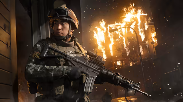A soldier in camouflage and a tactical vest with a scoped rifle stands at the edge of a wooden doorway; flames pour from the windows of a building across the street and sparks fill the air.