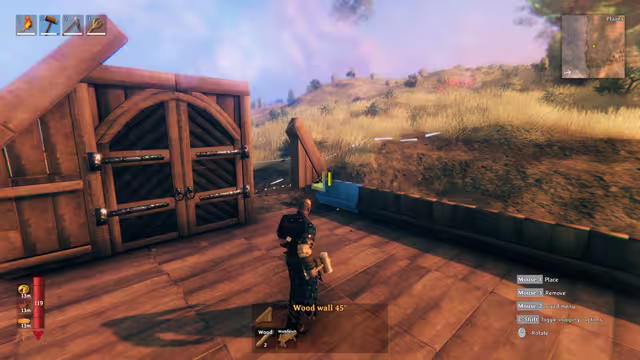 In-game screenshot of a Viking in dark armor holding a hammer on a wooden platform beside a large double barn door, with a translucent blue wall piece being placed ahead of him on yellow Plains grass; the hotbar, minimap and a 'Wood wall 45°' build panel are visible