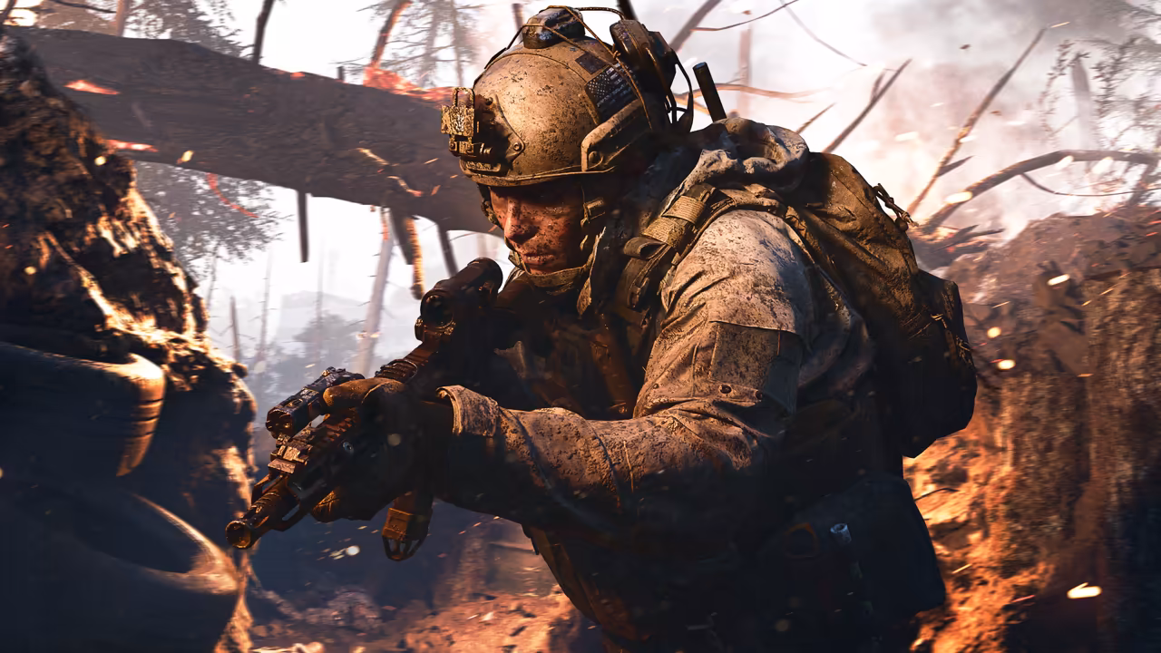 A soldier in a helmet and backpack, his uniform and face covered in brown mud, aims a rifle low in a dirt trench, with burning debris, splintered branches and smoke behind.