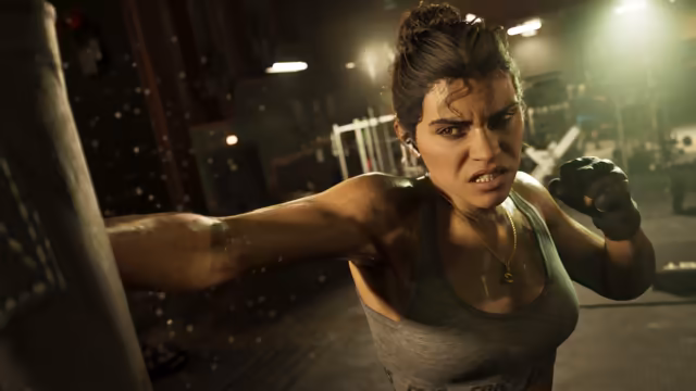 A woman with her hair tied up, wearing a gray tank top, fingerless gloves and earbuds, grimaces as she throws a punch at a hanging bag, sweat flying, in a dim gym with weight machines behind her.
