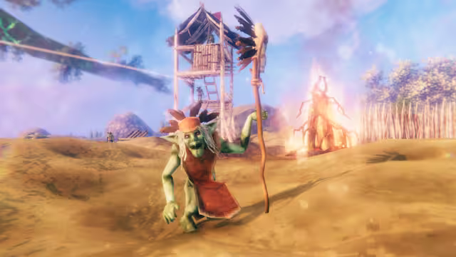 A green goblin in a red tunic and feathered cap hunches forward holding a tall staff topped with feathers; behind it stand a wooden watchtower, a burning pile of wooden spikes and a fence of sharpened stakes on sandy ground.