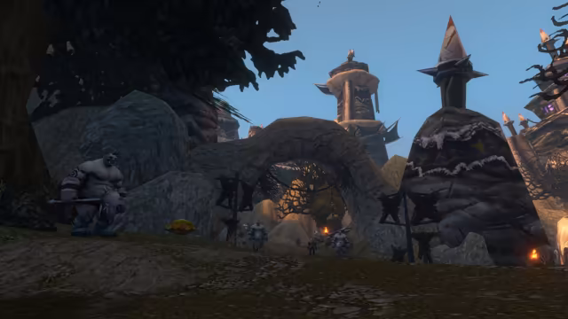 A gray-skinned ogre holding a spiked mace stands on a rocky rise at left, while more ogres gather on the path through a large rock arch flanked by dark banners on poles. Behind them rise tall towers with spiked roofs and fur trim, lit by torches against a pale blue sky.