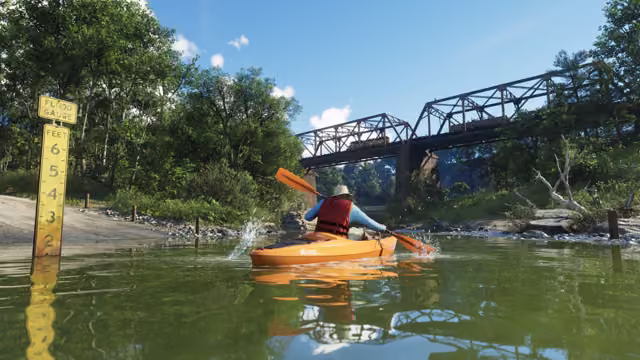 A person in a hat and a red life jacket paddles an orange kayak on a green river beside a yellow flood gauge marked in feet, with a steel truss railroad bridge and a thick forest behind.