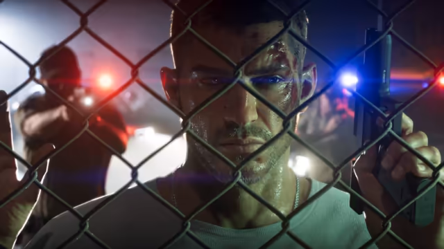 Close-up of a man with a cut forehead and a sweaty face looking through a chain-link fence, holding a pistol upright, while a figure aims a gun in the background amid red and blue police lights.