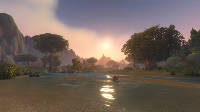 Low sunlight glints on a wide, shallow river with sandbanks, where a red crab-like creature stands in the water. Broad leafy trees, reeds and a red-roofed building line the banks, a scaly crocodile-like beast lies on the sand at the left, and jagged hills fade into a pink and purple sky.