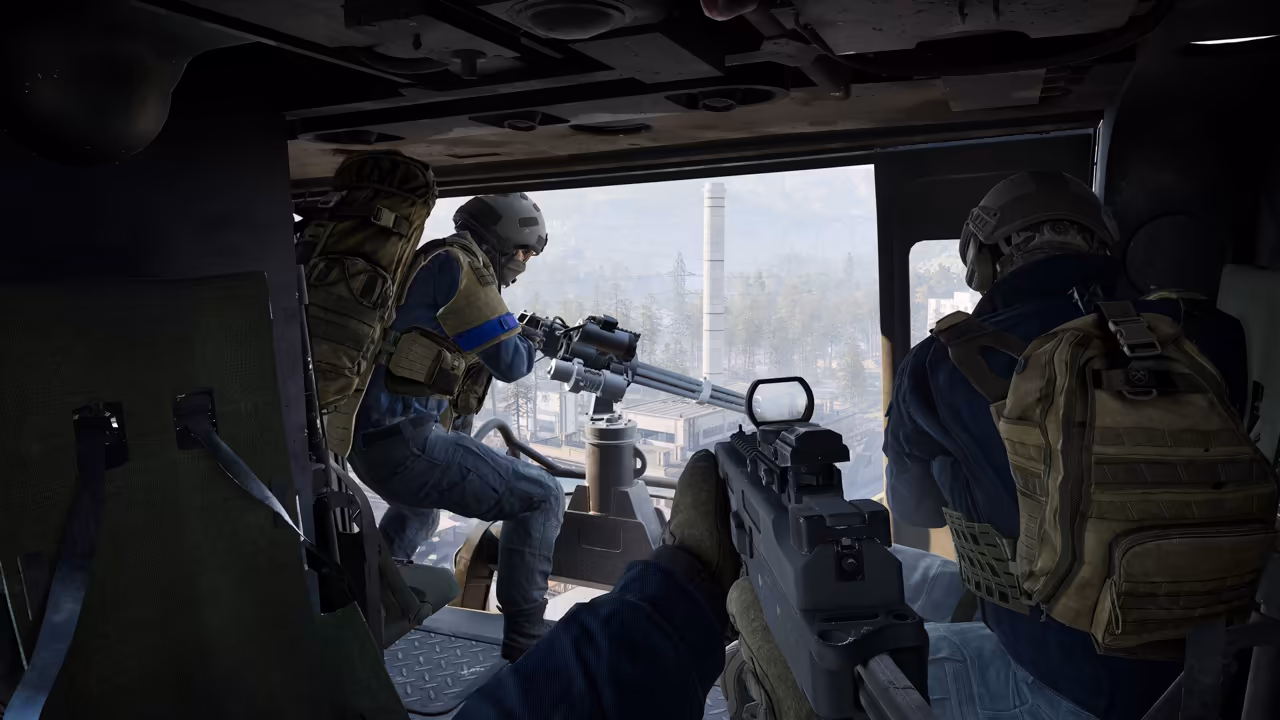 First-person view inside a helicopter's cabin, holding a rifle with a red-dot sight; a seated soldier in a blue armband mans a rotary gun at the open door, another soldier sits at right, and chimneys and snowy hills show outside.