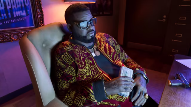 A bearded man in glasses, gold chains and an ornate red and gold jacket leans back in a cream armchair holding a thick wad of banknotes, a pistol on the desk beside him and a framed Jack of Hearts poster on the purple-lit wall.