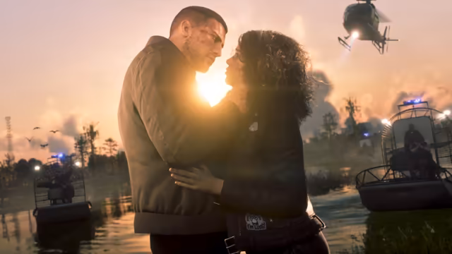 A man in a brown jacket and a woman with curly hair embrace, faces close, against a low sun over marsh water, while two airboats with flashing blue lights and a helicopter with a searchlight approach behind them.