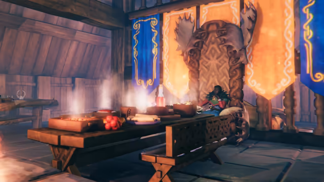 Screenshot of a timber hall: a long wooden table set with steaming dishes, rolled flatbread, a red bottle and a cluster of red fruit stands before a carved wooden throne topped with broad antlers, where a Viking in a green feathered cape and red sleeves sits with chin in hand; blue and orange banners with gold and white patterns hang behind, and a carved bench stands in front of the table