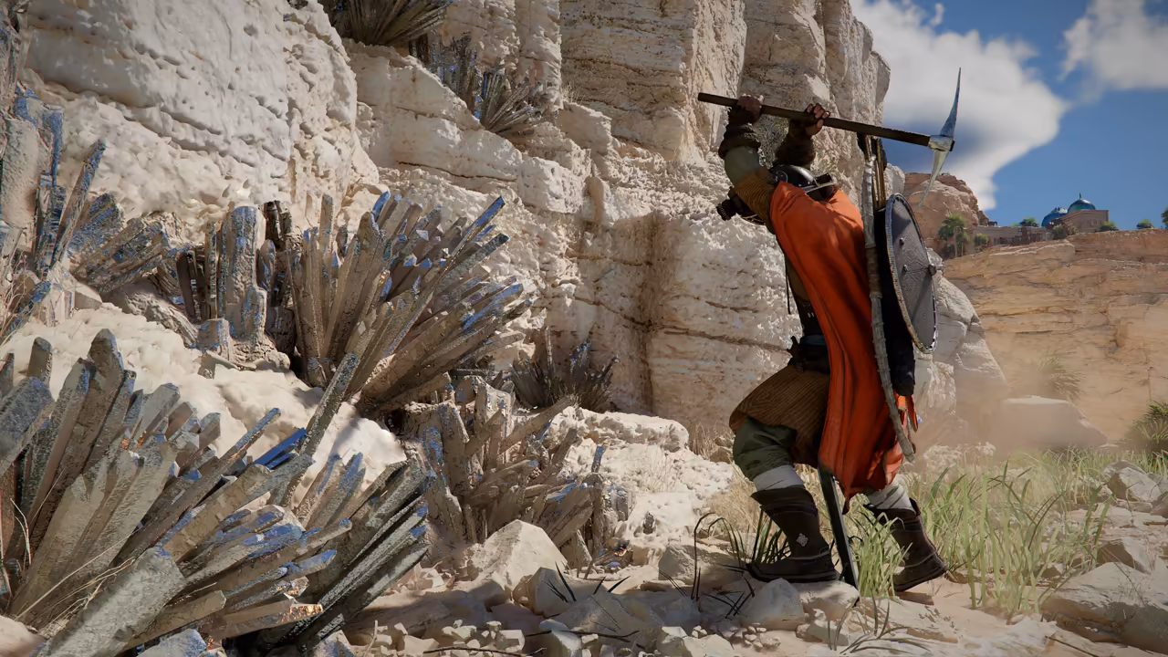 Against a white limestone cliff, a man in a bright orange cloak with a round shield on his back raises a pickaxe over his head to strike a cluster of long gray crystal spikes. Dry grass grows at his feet, and buildings with blue domes stand on the clifftop at right.